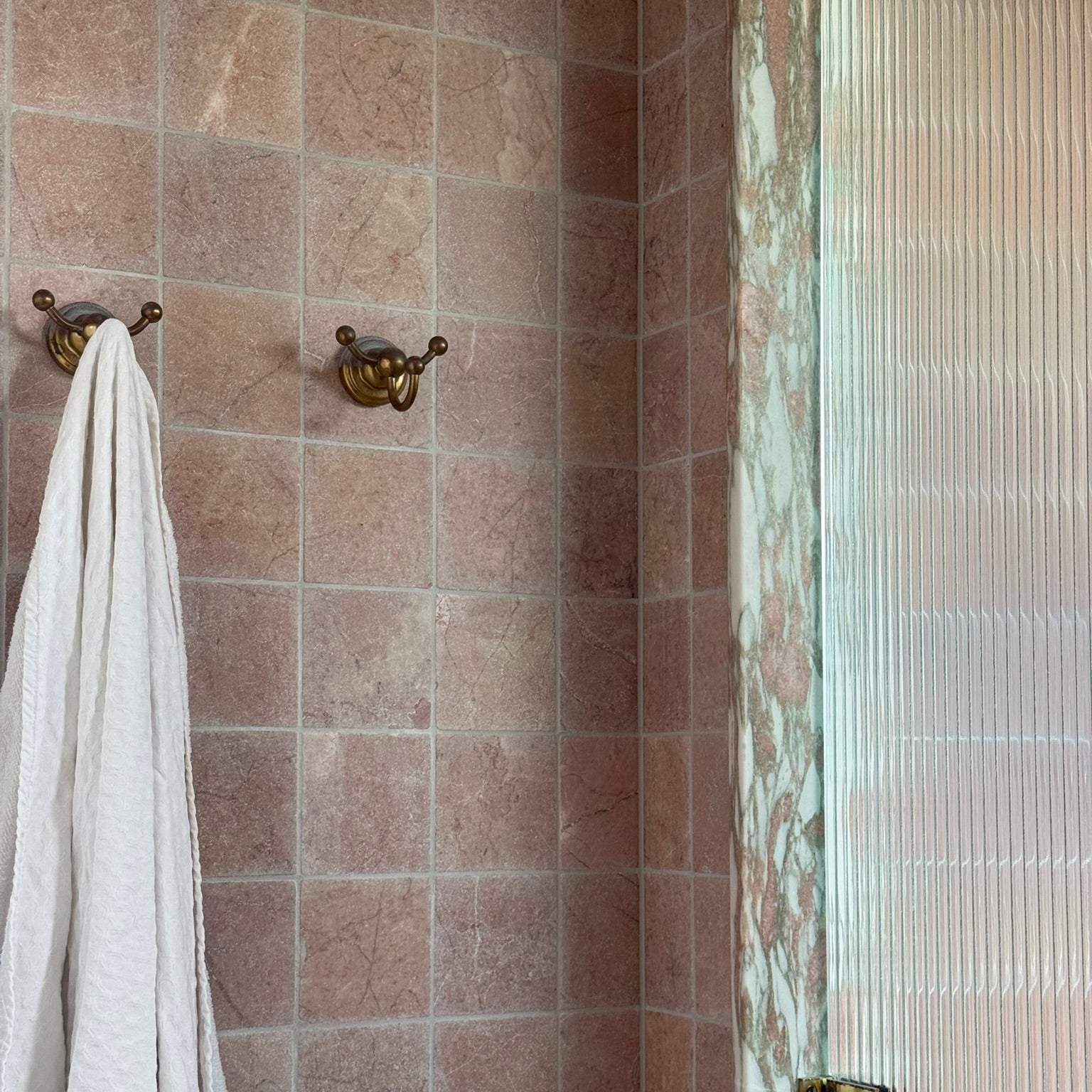 Towel hanging on a gold hook in a pink natural stone tiled bathroom corner with a glass shower door.